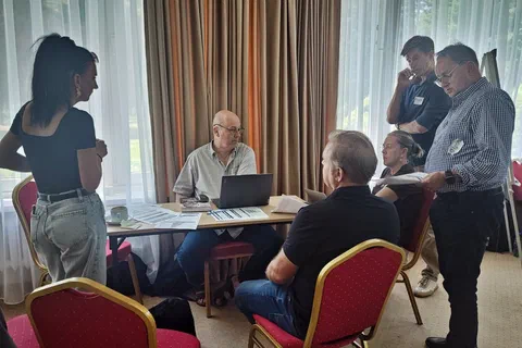 Zespół warsztatowy dyskutujący przy stoliku pod oknem sali szkoleniowej.