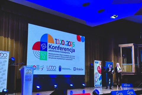 Scena auli konferencyjnej z dwiema prowadzącymi oraz dużym ekranem z tytułem i datą konferencji.
