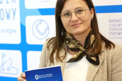 Uczestniczka konferencji w okularach prezentująca niebieską publikację na tle ścianki wydarzenia.