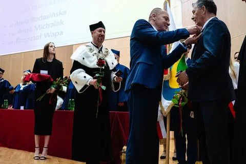 Przypinanie odznaczenia do marynarki wyróżnionego pracownika podczas uroczystości akademickiej w auli.