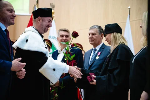Rektor w todze i birecie wręczający czerwoną różę i składający gratulacje na tle pocztu sztandarowego.