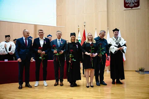 Wspólne zdjęcie odznaczonych osób z różami oraz władz uczelni w togach na tle pocztu sztandarowego i godła.