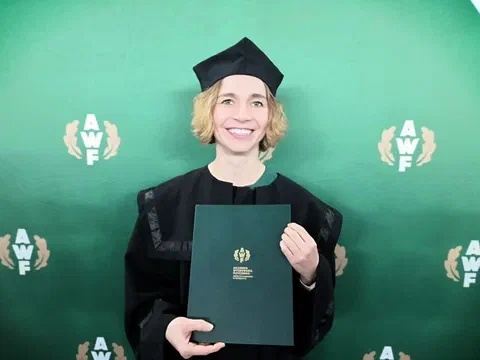 Dr hab. Dorota Sadowska w todze i birecie akademickim prezentuje dyplom habilitacyjny na tle ścianki AWF Warszawa