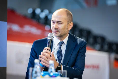 Prelegent w granatowym garniturze przemawiający do mikrofonu podczas debaty.
