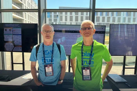 Dwaj badacze z identyfikatorami stojący przed monitorami podczas sesji e-posterowej.