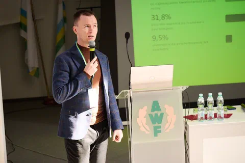 Prelegent w marynarce przemawiający przy mównicy na tle zielonego slajdu z wynikami procentowymi.