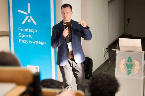 Prelegent z mikrofonem gestykulujący podczas wystąpienia obok baneru Fundacji Sportu Pozytywnego.
