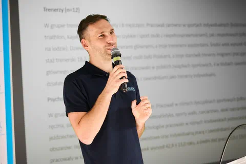 Zbliżenie na prelegenta z mikrofonem na tle slajdu opisującego badaną grupę trenerów i psychologów.