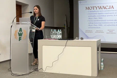 Prelegentka przy mównicy i stole prezydialnym obok ekranu ze slajdem poświęconym motywacji.