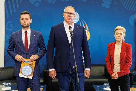Trzy osoby przemawiające na scenie na tle niebieskiej ścianki podczas otwarcia wydarzenia.