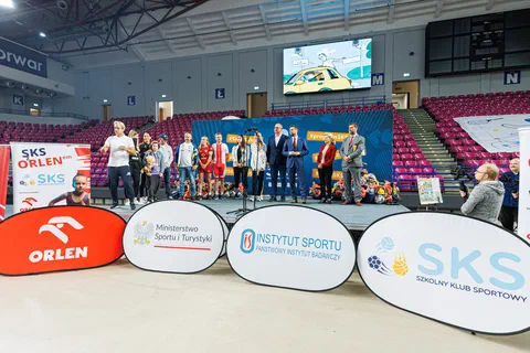 Grupa sportowców i organizatorów stojąca na scenie przed trybunami hali sportowej.