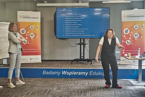 Dwie prowadzące omawiające slajd prezentacji o kompetencjach liderek przed ekranem i roll-upami Akademii Zarządzania Sportem.