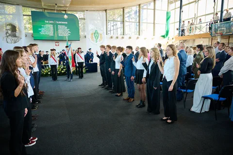 Studenci stojący w rzędach w hali podczas uroczystego ślubowania akademickiego przed pocztem sztandarowym.