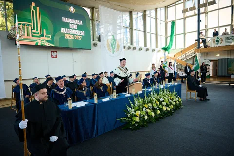 Władze uczelni w togach stojące za stołem prezydialnym udekorowanym kwiatami na tle baneru inauguracyjnego.