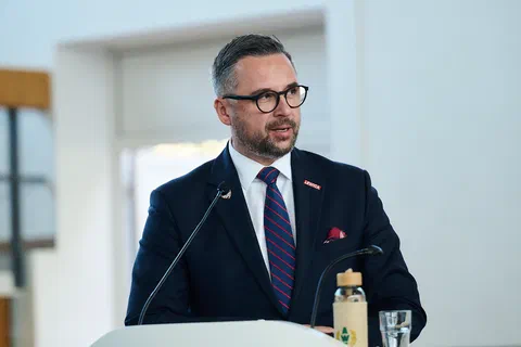 Mężczyzna w granatowym garniturze i okularach przemawiający do mikrofonu z mównicy.