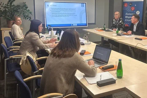 Uczestnicy spotkania projektowego siedzący przy stołach z laptopami podczas prezentacji przy ekranie.