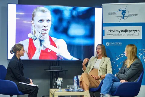 Trzy uczestniczki rozmowy siedzące w fotelach przed ekranem ze zdjęciem lekkoatletki i banerem Akademii Trenerskiej.