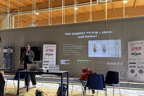 Prelegent występujący przy stoliku z laptopem przed ekranem projekcyjnym w hali szkoleniowej.