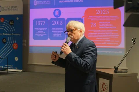 Prelegent w okularach omawiający z mównicy slajd z historią i statystykami na lata 1977–2025.