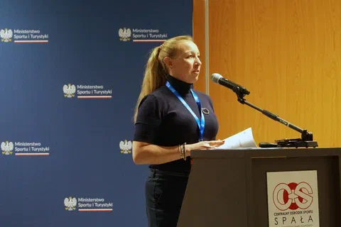 Kobieta występująca przy mównicy z logo Centralnego Ośrodka Sportu w Spale.