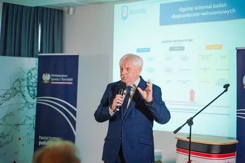 Prelegent w garniturze przemawiający do mikrofonu na tle ekranu ze schematem badań diagnostyczno-wdrożeniowych.