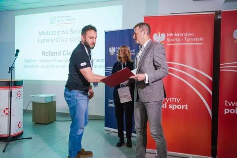Wręczenie dyplomu w bordowej oprawie trenerowi na tle banerów Ministerstwa Sportu i Turystyki.