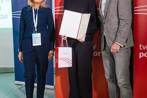 Trzy osoby pozujące z wręczonym dyplomem na tle czerwono-niebieskich banerów resortu.