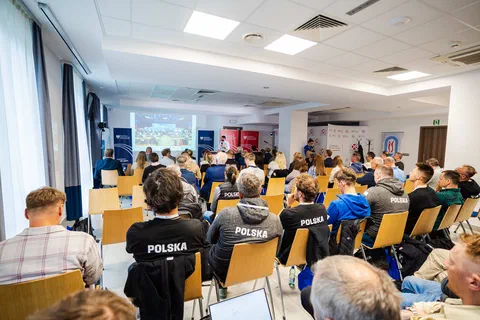 Widok z tyłu sali na uczestników konferencji w koszulkach z napisem „Polska”.