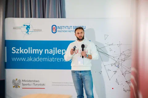 Prelegent z mikrofonem występujący na tle ścianki Akademii Trenerskiej i Instytutu Sportu – PIB.