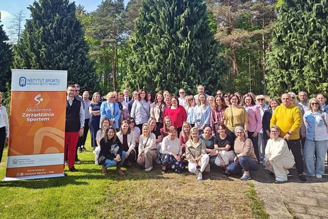 Grupowe zdjęcie uczestników szkolenia w plenerze na tle zieleni i baneru Akademii Zarządzania Sportem.