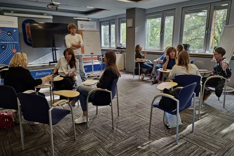 Uczestniczki warsztatów rozmawiające w parach i małych grupach na krzesłach z pulpitami w sali.
