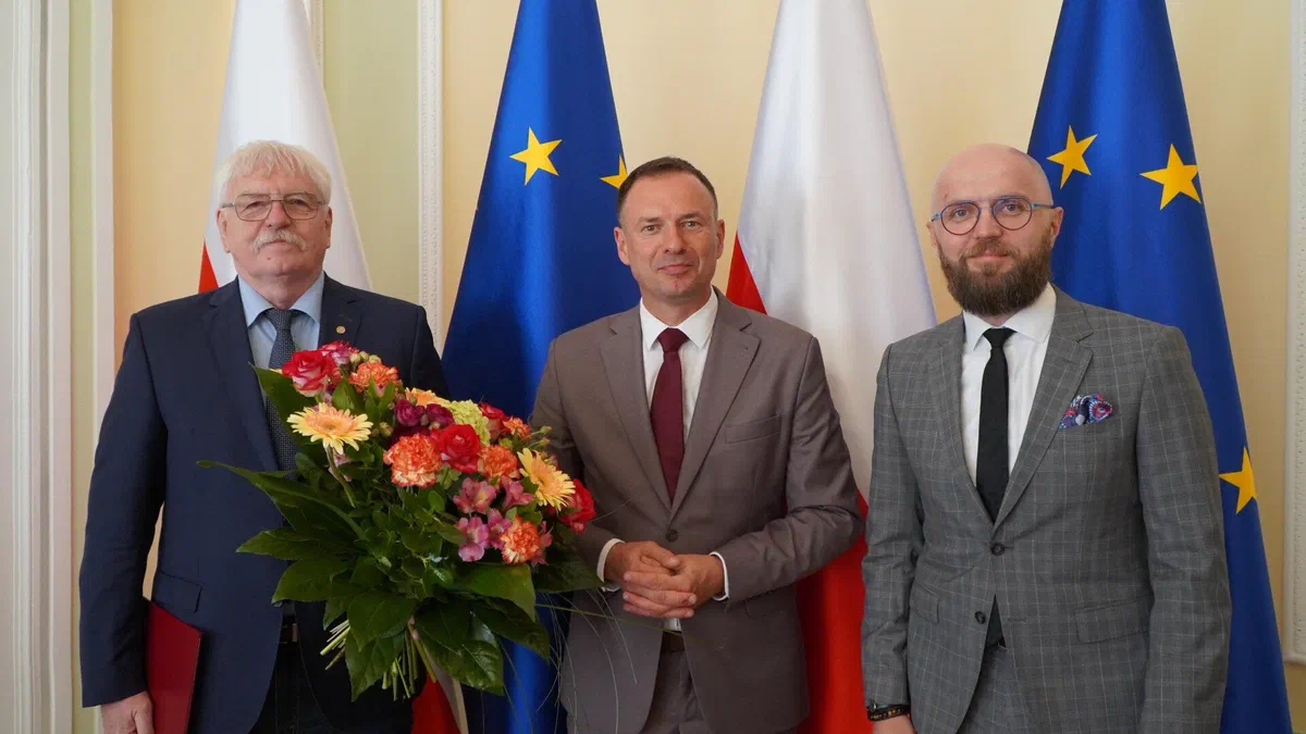 Trzej mężczyźni na tle flag Polski i Unii Europejskiej podczas uroczystości objęcia stanowiska Dyrektora Instytutu Sportu – PIB przez Marka Droba; ustępujący dyrektor dr Jerzy Eliasz trzyma bukiet kwiatów.