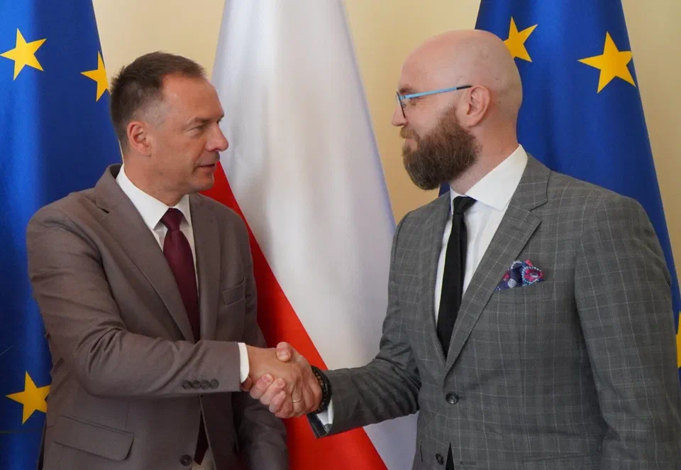 Uścisk dłoni podczas przekazania obowiązków Dyrektora Instytutu Sportu – Państwowego Instytutu Badawczego Markowi Drobowi na tle flag państwowych i unijnych