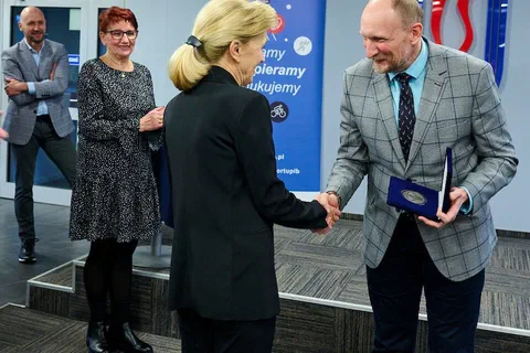 Wręczenie medalu w otwartym etui i uścisk dłoni na tle ścianki z logo Instytutu Sportu