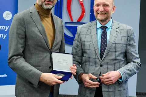 Dwaj mężczyźni w marynarkach pozujący z przyznanym medalem w etui przed logo Instytutu