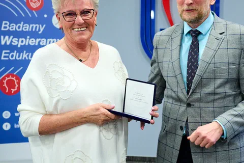 Kobieta w jasnej bluzce prezentująca medal w etui obok wręczającego podczas uroczystości