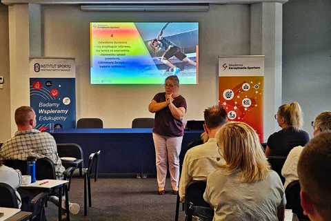 Prowadząca stojąca przed stołem prezydialnym i omawiająca slajd prezentacji przed uczestnikami