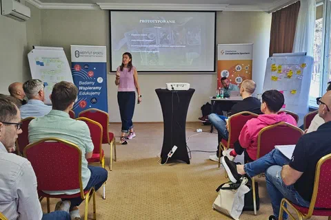 Uczestniczka prezentująca na środku sali obok ekranu z hasłem Prototypowanie i flipchartów