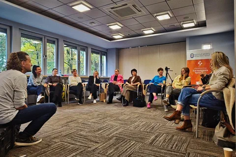 Uczestniczki siedzące w półkolu pod oknami i słuchające prowadzącego siedzącego po prawej stronie