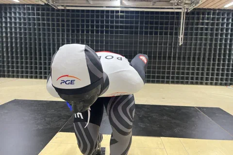 Łyżwiarz szybki w kasku i kombinezonie reprezentacji Polski ustawiony w niskiej pozycji w tunelu aerodynamicznym