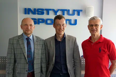 Trzej mężczyźni pozujący na tle ściany z przestrzennym niebieskim napisem Instytut Sportu