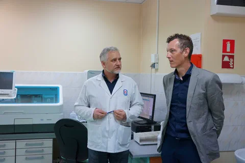 Pracownik laboratorium w białym fartuchu objaśniający działanie aparatury analitycznej gościowi w marynarce