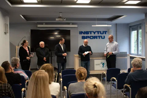 Pięciu mężczyzn stojących z przodu sali i przemawiających do uczestników wernisażu wystawy