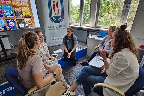 Cztery uczestniczki dyskutujące w podgrupie obok roll-upu Instytutu Sportu i tablicy z kartami