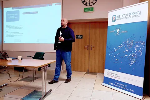 Wykładowca przemawiający przez mikrofon obok ekranu projekcyjnego i niebieskiego roll-upu Instytutu Sportu