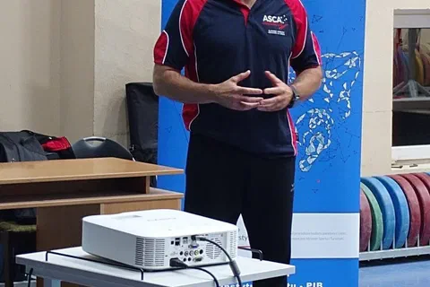 Prelegent w stroju sportowym przemawiający na tle roll-upu przed projektorem