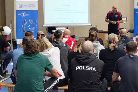 Wykładowca w stroju sportowym omawiający schemat na ekranie przed uczestnikami siedzącymi w sali