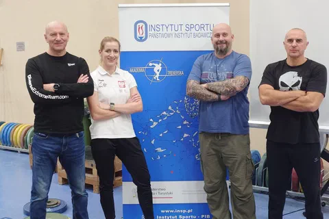 Czworo prowadzących w strojach sportowych pozujących obok roll-upu Instytutu Sportu w sali ze sprzętem ciężarowym