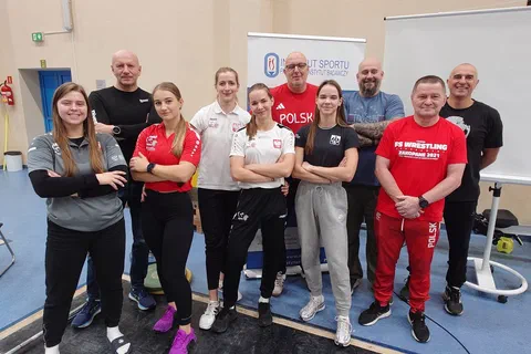 Grupa ośmiorga uczestników i prowadzących w strojach treningowych pozująca w sali ćwiczeń obok sztangi i tablicy