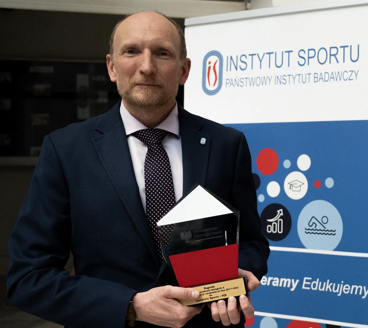 Dyrektor Instytutu Sportu – PIB dr Konrad Witek trzymający statuetkę przyznaną podczas Kongresu „Nauka dla Społeczeństwa” na tle roll-upu Instytutu.